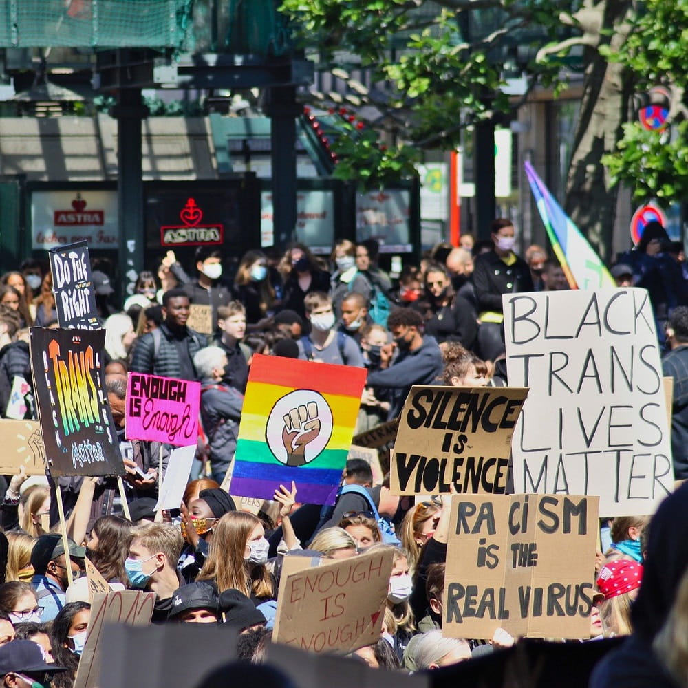 An outdoor protest with people holding placards, one reading "Black Trans Lives Matter", "Racism is the real virus", and "Silence is violence"