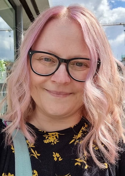 Essex graduate Carolyn White, with pink hair and glasses, standing outside with a window, blue sky and clouds in the background.