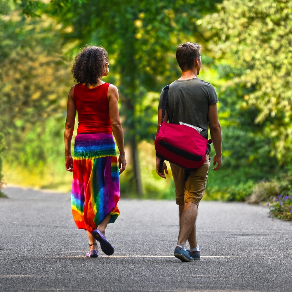 A woman on the left and a man on the right walking down a tree-lined path, with woman is wearing a rainbow-coloured skirt.
