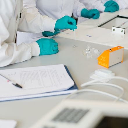 A group of people, heads out of shot, wearing white lab coats and green disposable gloves, working at a lab bench with a notebook, petri dish and little glass bottle. One of the hands is holding a swap and another is holding a microscope slide.