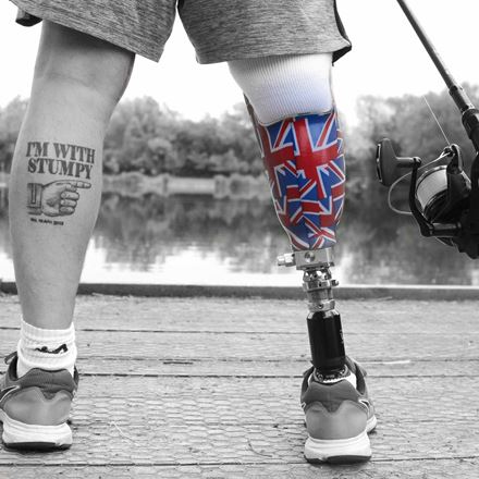 Someone wearing shorts standing in front of a body of water. Their right leg is a prosthetic leg with a Union Jack pattern, and their left leg has a tattoo of a hand pointing and the words "I'm with stumpy".