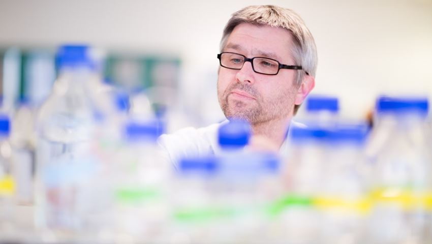 Dr Ralf Zwacka looking over a collection of clear bottles with blue lids and some green and yellow labels.