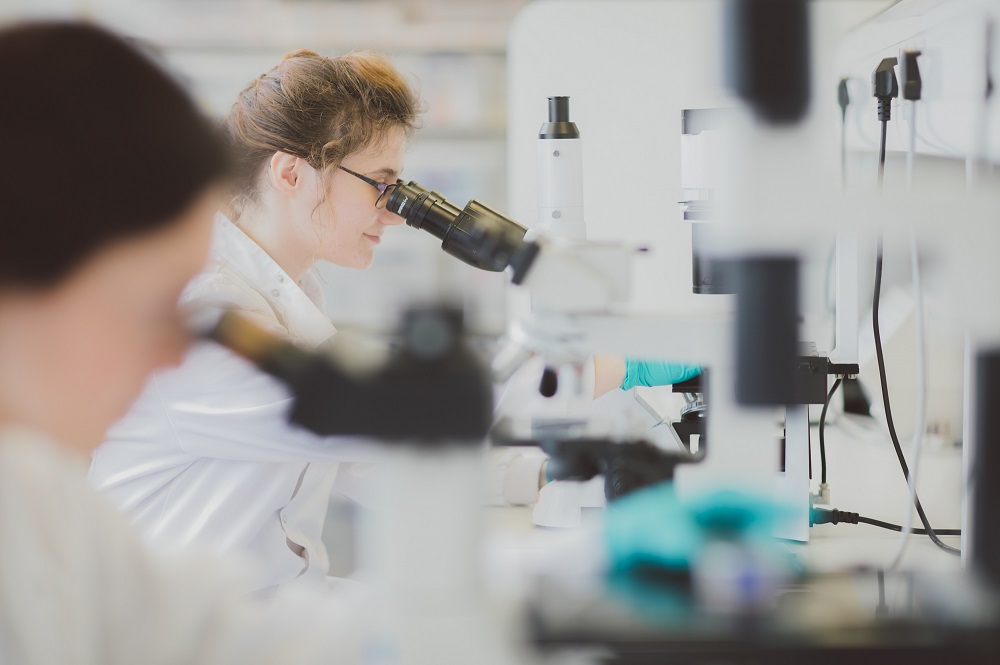 Two people, a blurry one in the foreground with the focus on someone in the background, looking down microscopes in a lab.