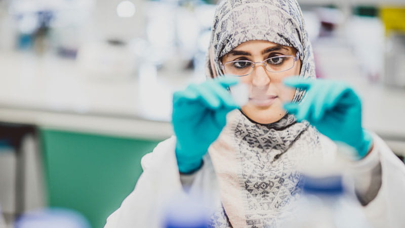 A woman wearing a headscarf, a lab coat and green gloves, holding up a microscope slide.