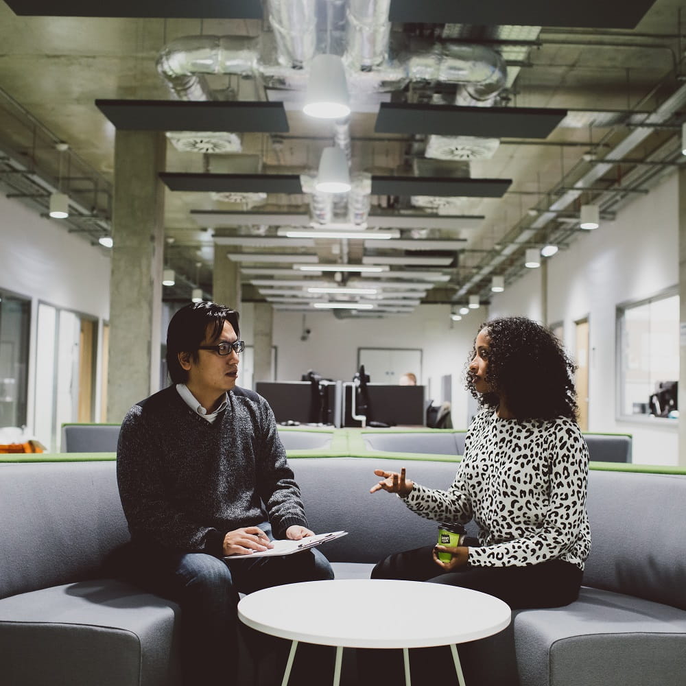 Two people sitting on a large grey sofa in the Department of Mathematical Sciences, one gesturing as she talks.