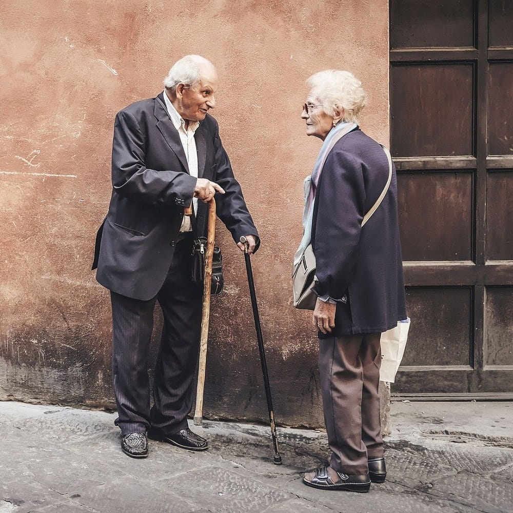 Two elderly people standing in front of a terracotta-coloured wall, talking together.