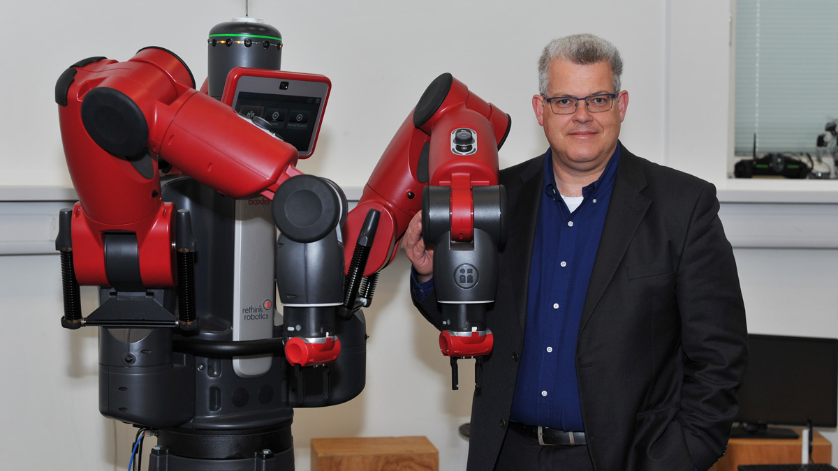 A photo of Professor Klaus McDonald-Maier, from the School of Computer Science and Electronic Engineering, standing next to a large red and black Baxter robot.)