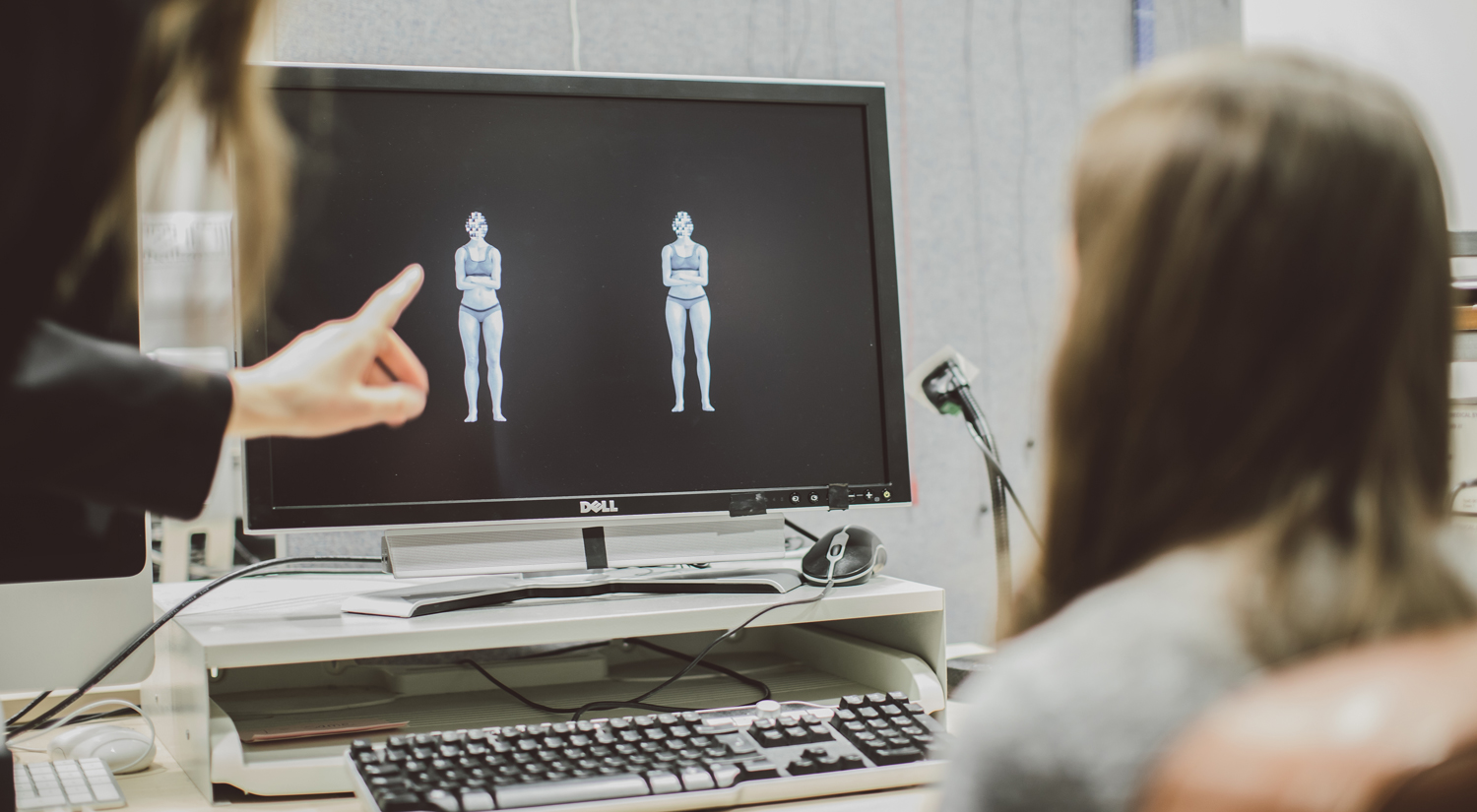 In the centre of the photo is a computer screen showing two computer-generated figures in white on a black background. To the right of the photo is a student sitting with their back to the camera. On the left, mostly out of shot, is a second person pointing at the screen with their left hand and index finger.