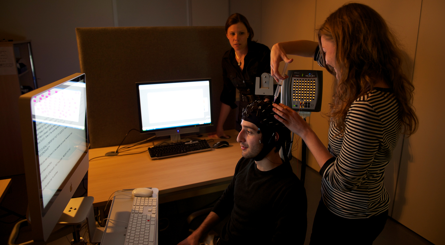 A student wearing an EEG cap is sitting in front of a large computer monitor. A second student is standing next to them, injecting some gel in to one of the cap sensors. Professor Silke Paulmann from the Department of Psychology is standing in the background, next to a second computer, watching them.
