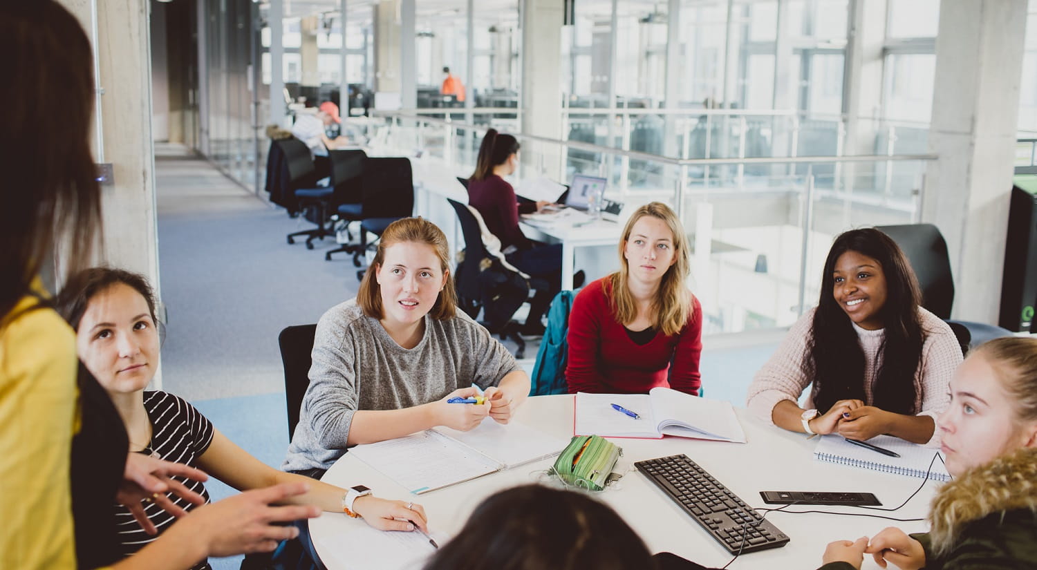 A group of students sitting at a large round table together, looking towards the speaker in front of the camera.