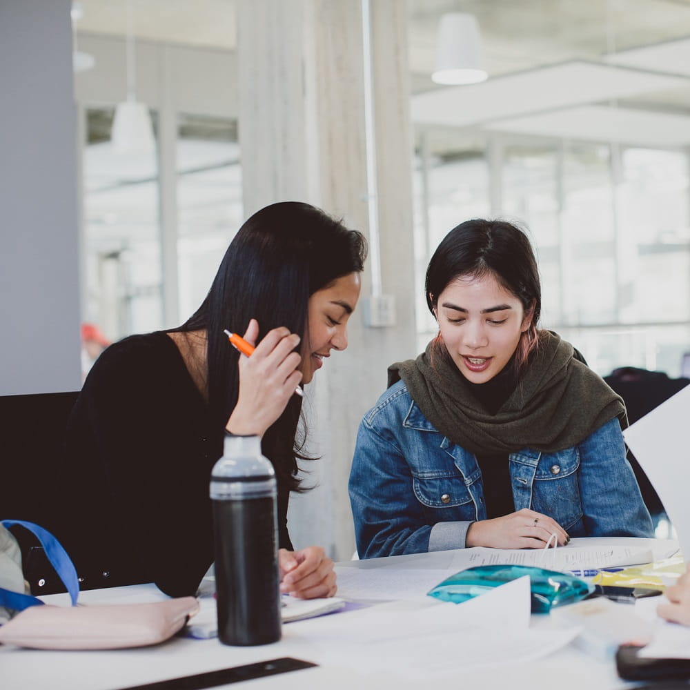 Two young women working at a table covered in papers, pencil cases and a water bottle. The one on the left is leaning towards the one on the right to look at a piece of paper on the table.