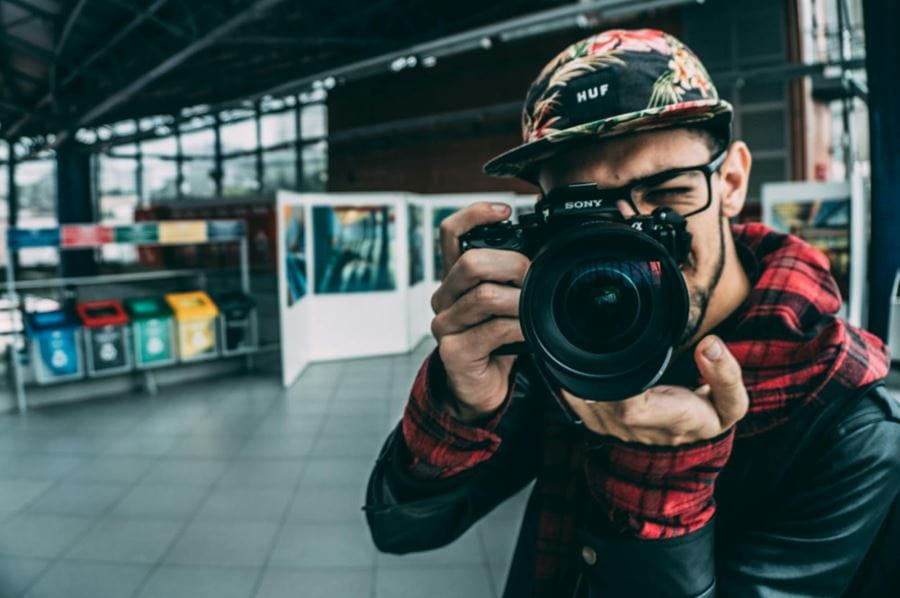 A man standing in a building, holding a camera and looking through it at the photographer taking this photo.