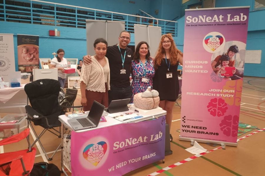 Four PhD students standing in a school sports hall, with a table and a large pink and purple banner advertising the SoNeAt Lab.