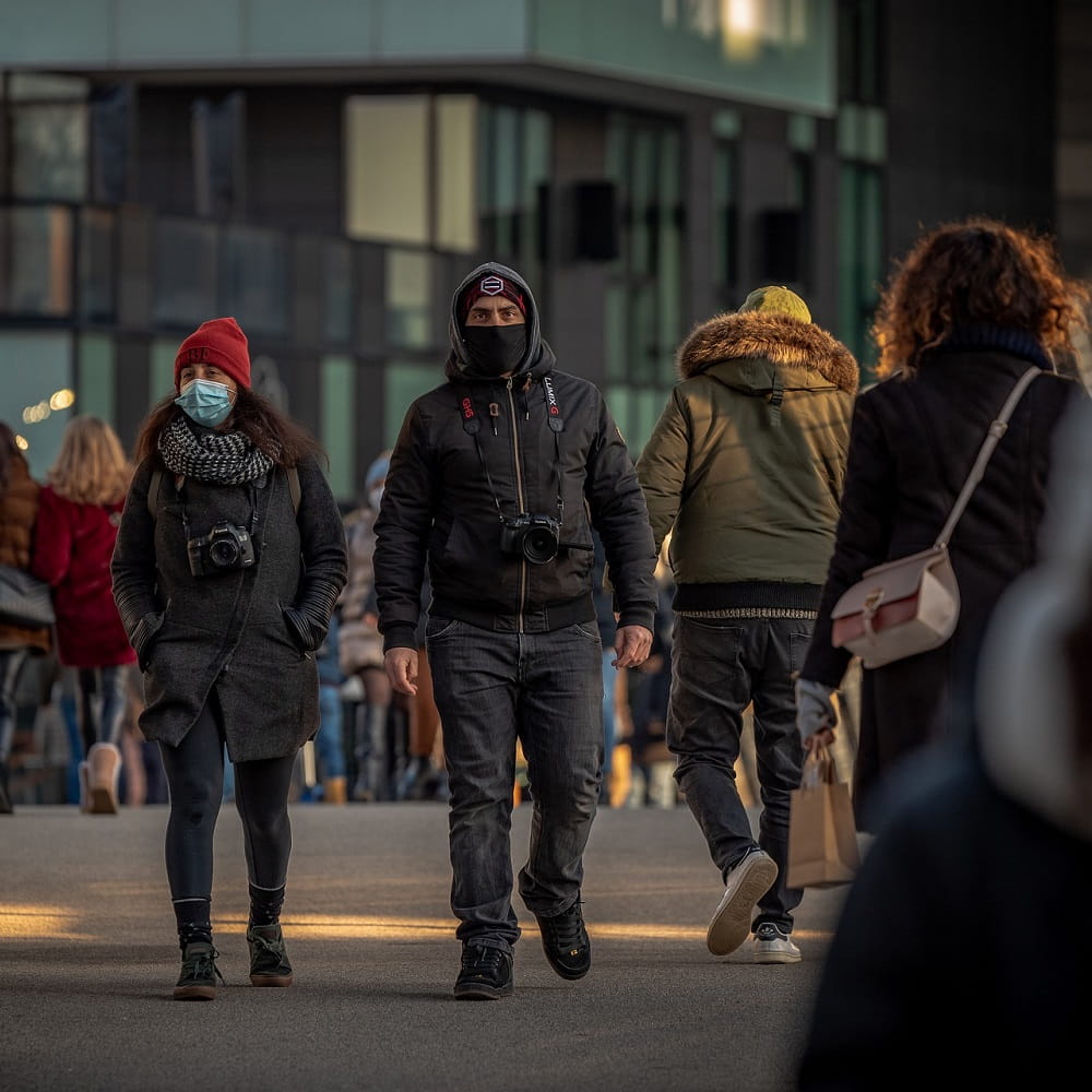 Two people wearing face mask walking towards the camera, with people walking past them in the opposite direction.