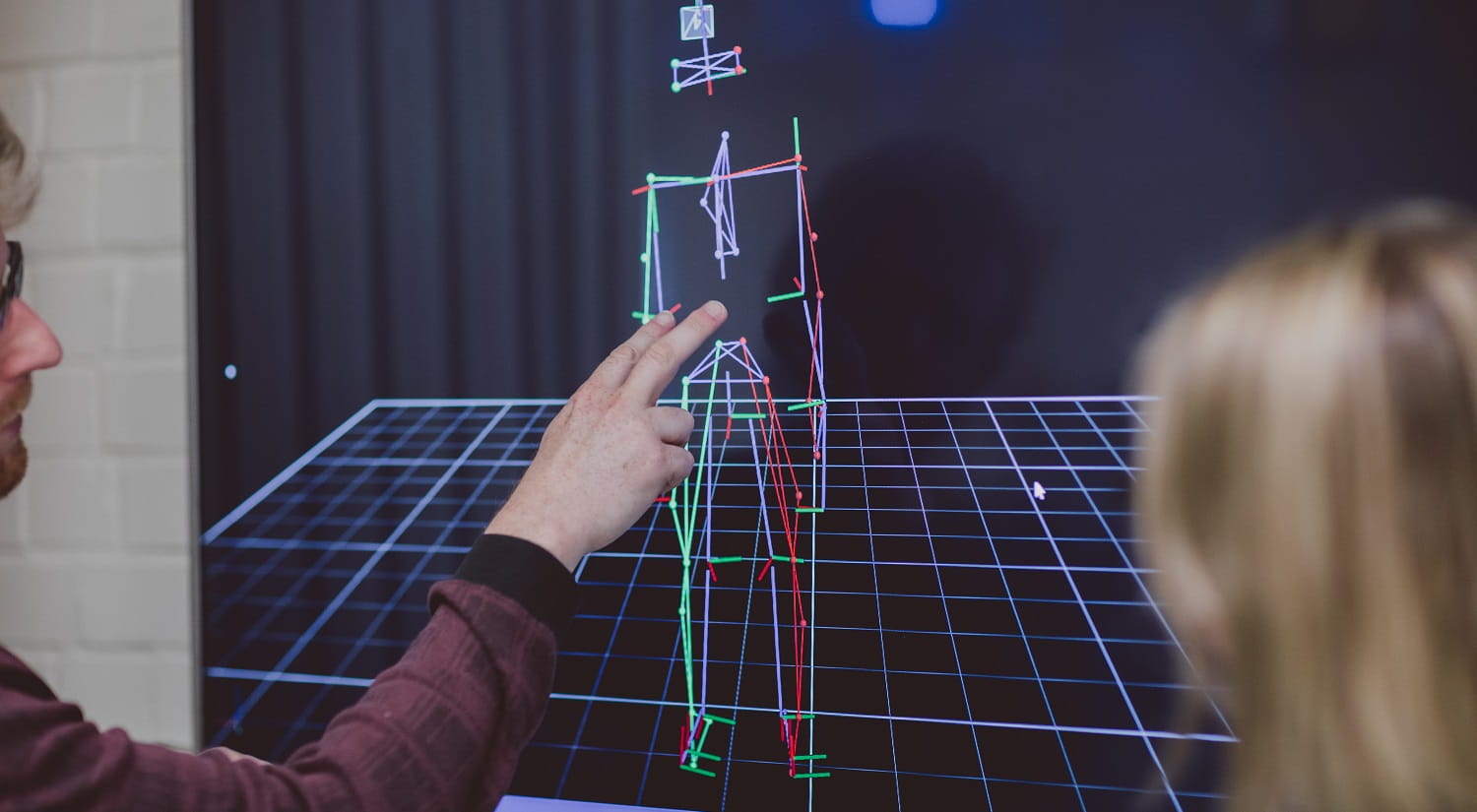A Sport, Rehabilitation and Exercise Sciences lecturer is mostly out of shot on the left. His hand is pointing at a screen with a stick figure in several colours, depicting an athlete recorded with the Vicon Motion Analysis system. The back of a student's head is in the foreground on the right.