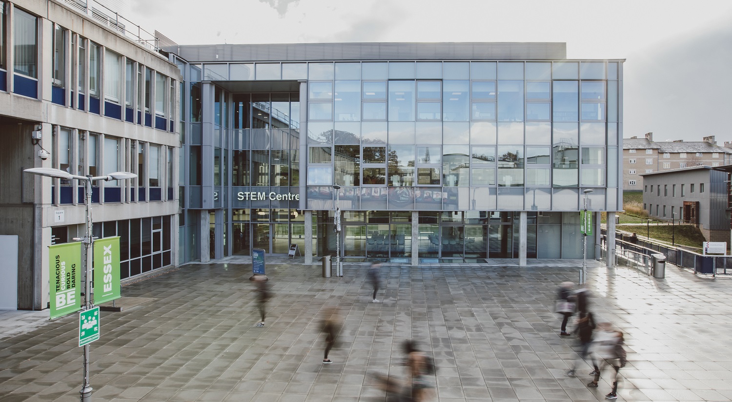 A long-exposure shot of the front of the STEM Centre on Colchester campus. A series of blurry figures are crossing the square in front, going in different directions.