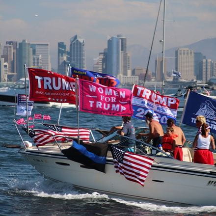 A private boat covered in flags supporting Donald Trump, being sailed across a lake with a city skyline in the background.