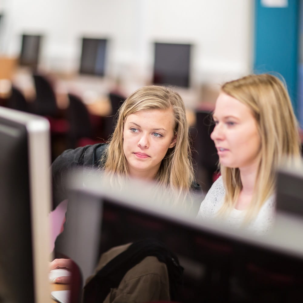 Two female students are sitting at a computer in a PC lab, with the student one the left leaning to the right slightly to get a better view of the screen.