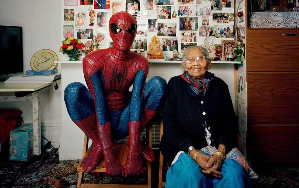 Hetain Patel in a full Spiderman outfit, crouched on a chair while in a Spiderman pose. Next to Hetain is his grandmother, they are both sat in her home, side by side and facing the camera.