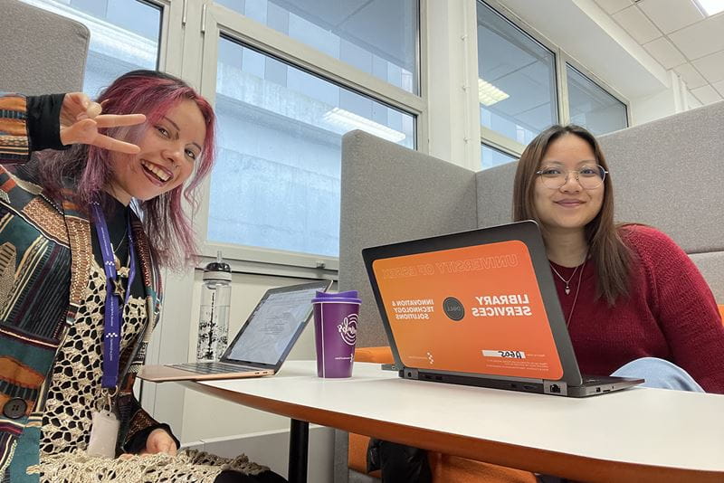 Sofia and Mimi sat at opposites sides of a desk, both resting laptops on the shared table. Both are looking towards the camera and smiling. 