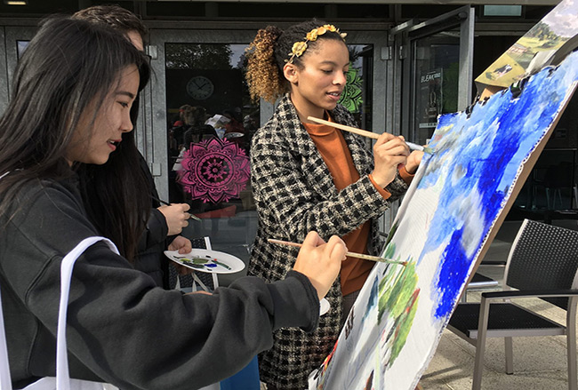 Three people, with paintbrushes in hand, painting Wivenhoe Park on an easel. Trees and sky has been painted onto the canvas but the painting is still a work in progress.
