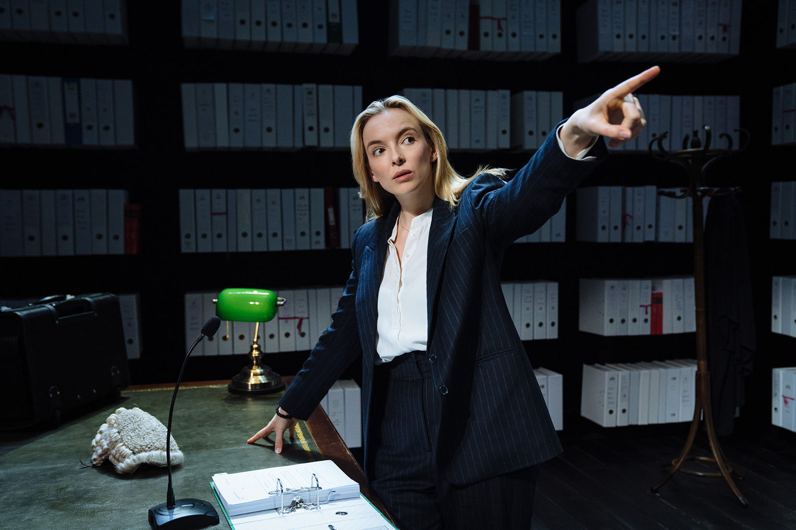 Jodie Comer as Tessa in Prima Facie. Tessa is dressed smartly in a suit, and is pointing off into the distance. Tessa's other hand is leaning against a green leather topped desk, and there are shelves of files behind her.