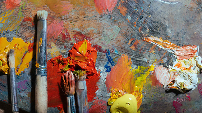 Paintbrushes of all different sizes, resting on a well-loved painting palette which is streaked with colourful dried paint.
