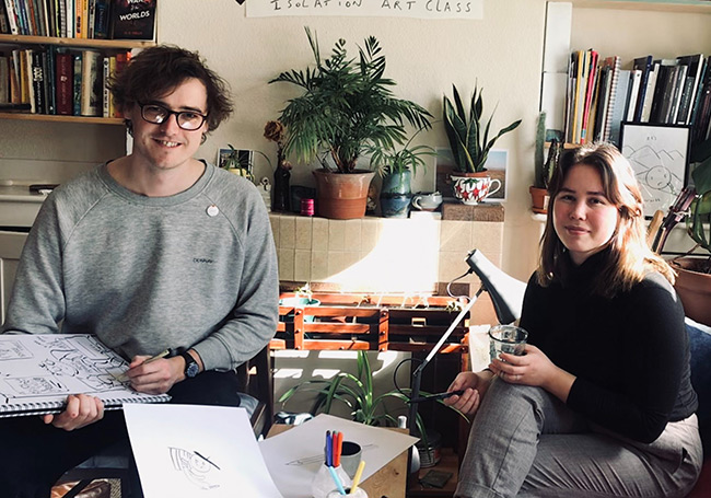 Artists Tom and Iris are sat in a living room, smiling towards the camera. Tom is holding a large sketchbook which Tom is in the process of drawing on.