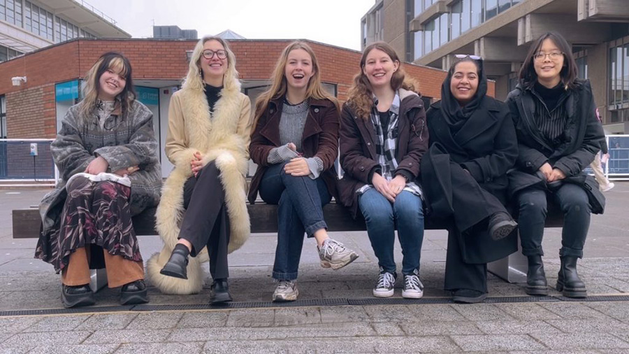 The six MA Curating and MA Museums and Heritage students sitting in a line on a bench together outside Art Exchange all smiling at the camera. LTR: Vendija Zile, Carla Reboud, Scarlett Angove, Lauren Wainwright, Freeha Hashmi, and Yu Li.