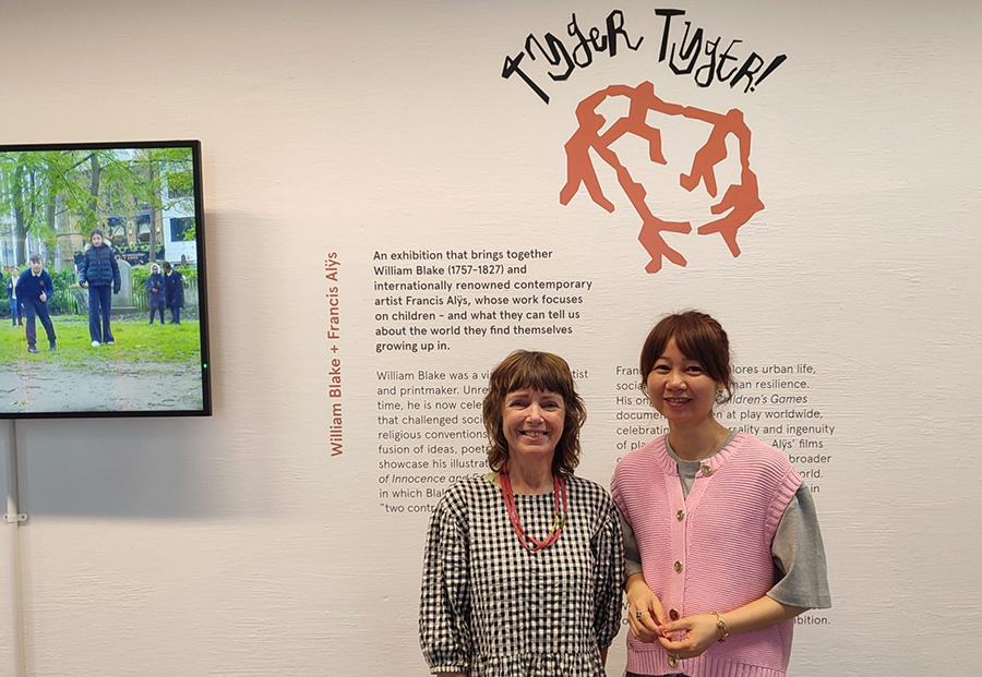 Jess Twyman and Anna Li stood in front of the wall text of the exhibition 'Tyger Tyger!', and smiling towards the camera.