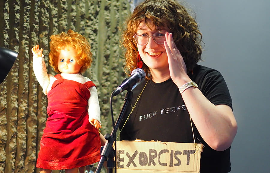 Alice K Stephens performing in front of a microphone, at a Lakeside Theatre Scratch Night event. Alice is holding a porcelain doll up in the air, and has a cardboard sign around her neck (Alice, not the doll). 