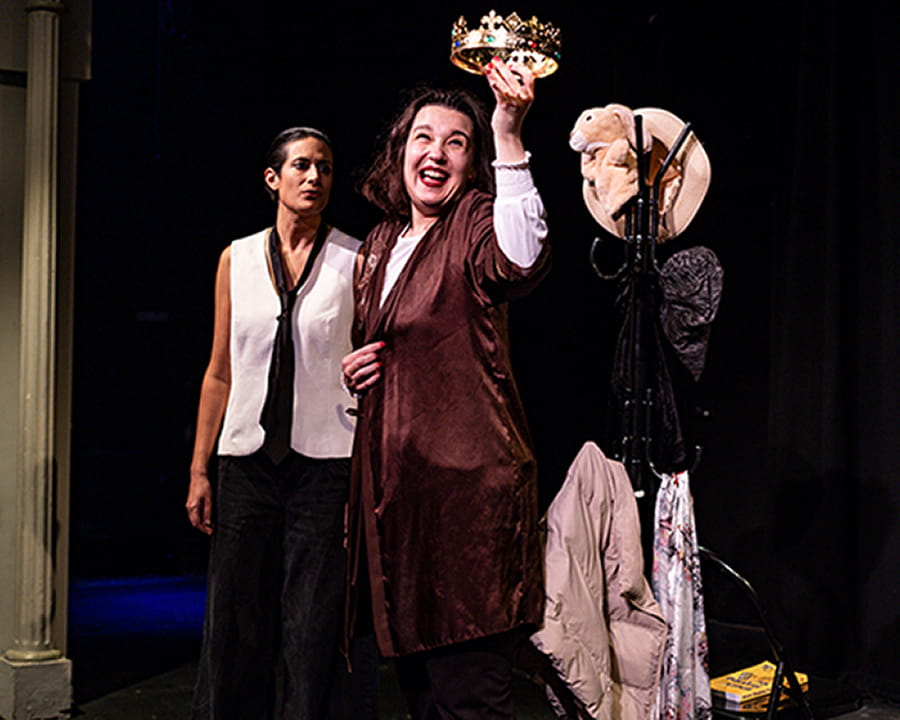 Performance shot of A Two Woman Hamlet. Two women on stage, with one holding a crown up into the air. 