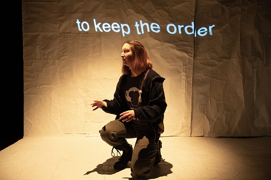 Marina Cusí Sanchez performing on stage during a performance of 'Sub Titles Over'. Behind Marina, the words 'to keep the order' are projected on a backdrop.