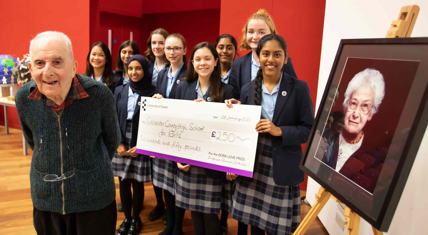 Girls from the Colchester County High School for Girls smiling and holding a large, over-sized cheque, with Holocaust survivor Frank Bright MBE pictured, smiling, in the foreground