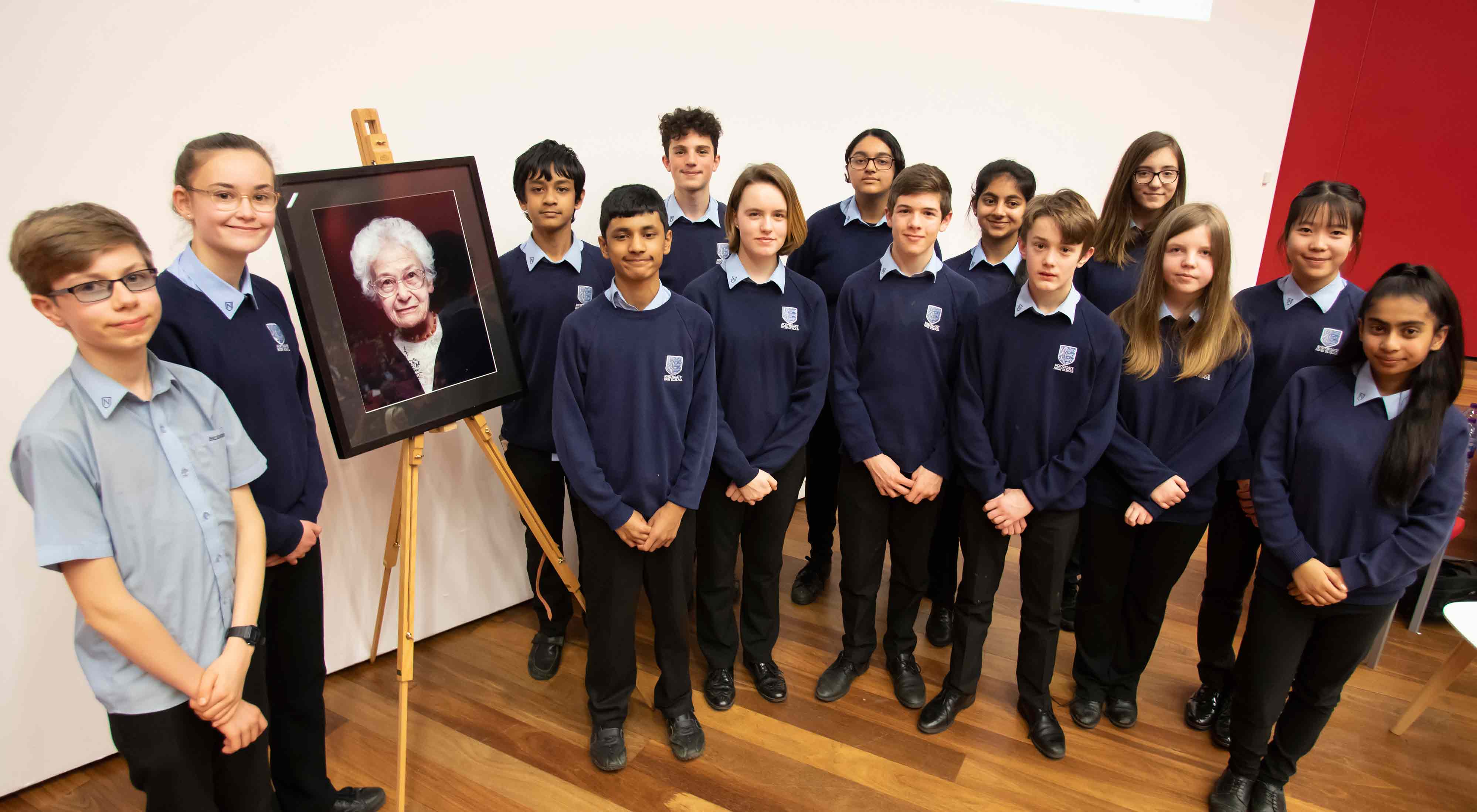A group of young children, smiling and looking relaxed, stand around the portrait of Holocaust survivor Dora Love which is resting on an easel