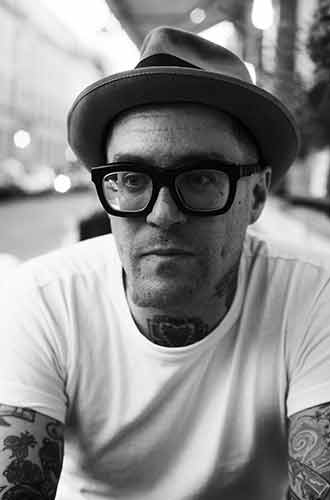 Dr Matt Lodder, photographed in black and white wearing a white t-shirt, black-rimmed glasses and a trilby-style hat