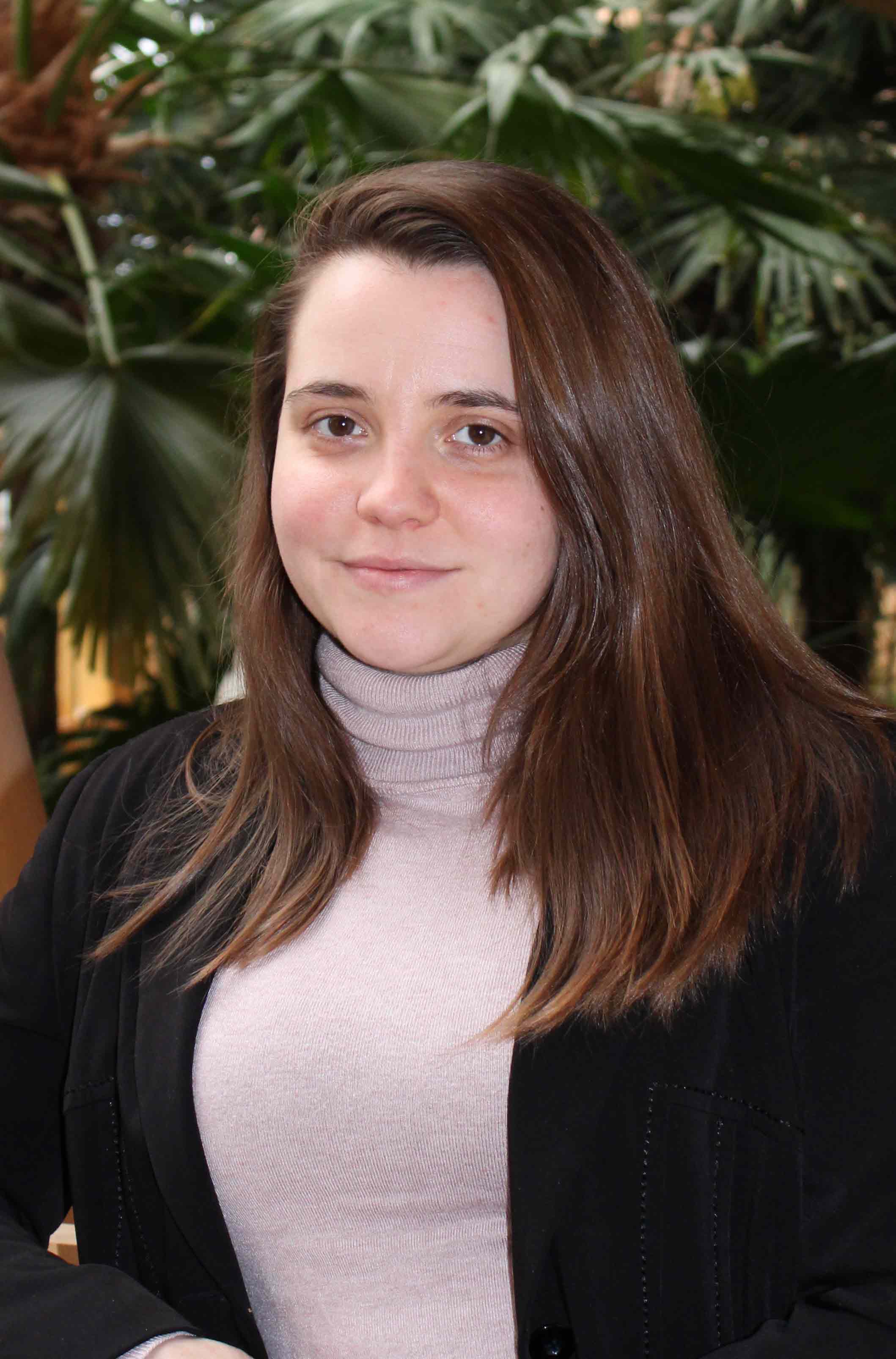 Dr Rebecca Warren, smiling and wearing a pink roll-neck top and black jacket, in front of a green plant