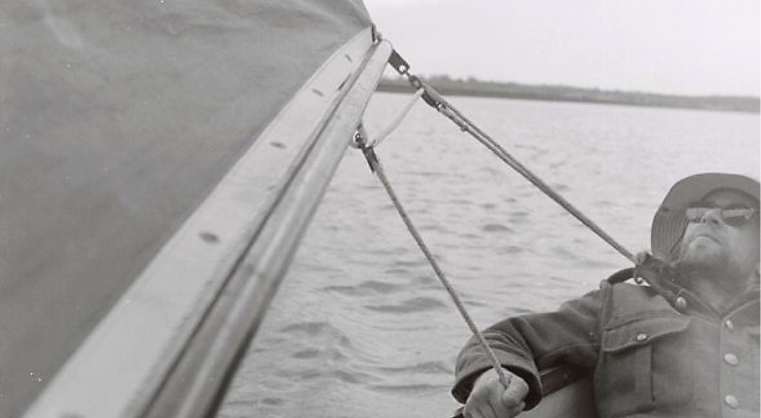 A man, wearing sunglasses, coat and hat, leans back in the bow of a boat with the sail flapping next to him