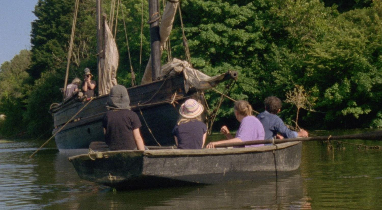 A group of people, sitting in a rowing boat on a sunny river watch as a barge approaches