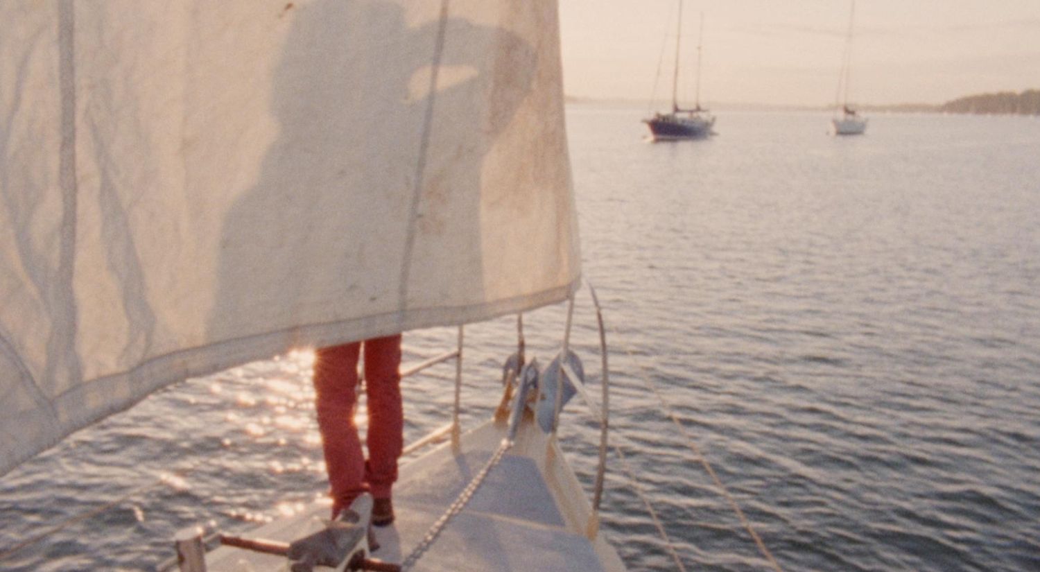 A person is seen in shadow behind a sail, standing on the deck of a boat, at sunset