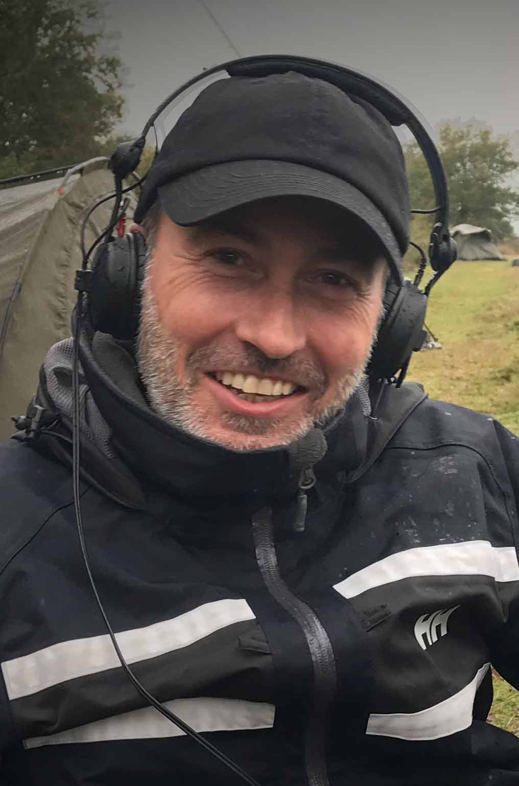 Nic Blower, smiling, wearing a black jacket, black baseball cap and headphones