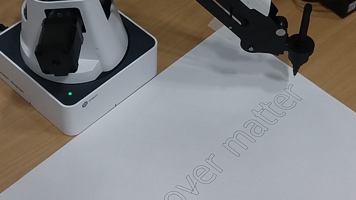 A robot arm holding a pen over a piece of paper, the words "over matter" are written in black.