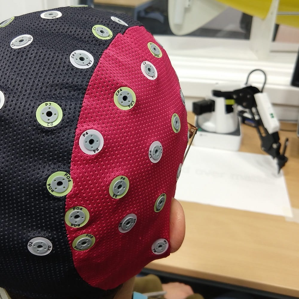 The back of the head of someone wearing a black and red EEG cap. Out of focus in the background is a robotic arm holding a pen over a piece of paper.