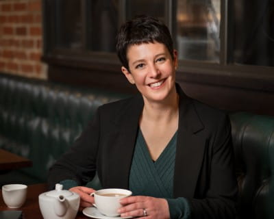 Charlotte Noon sitting at a table holding a cup, with a teapot in front of her.