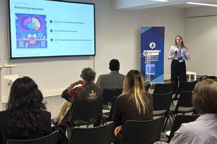 Five people with their backs to the camera, looking at Dr Raysa Rocha deliver her talk. There is a screen on the wall with a talk slide in the background.