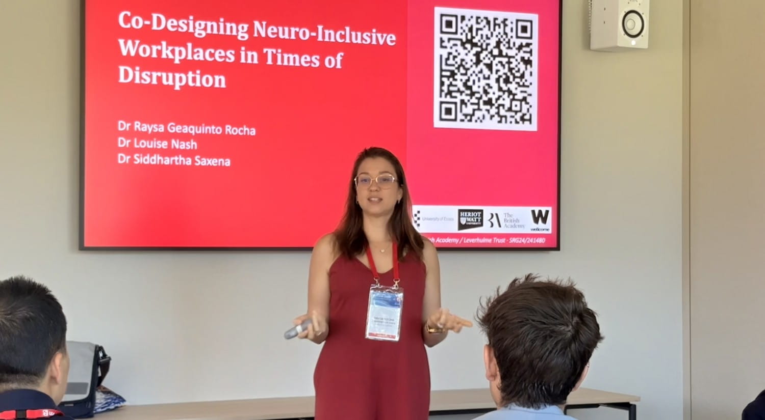 Dr Raysa Rocha standing at the front of a room with a few people in the foreground. A slide with the title "Co-Designing Neuro-Inckusive workplaces in times of disruption" is on the screen behind her.