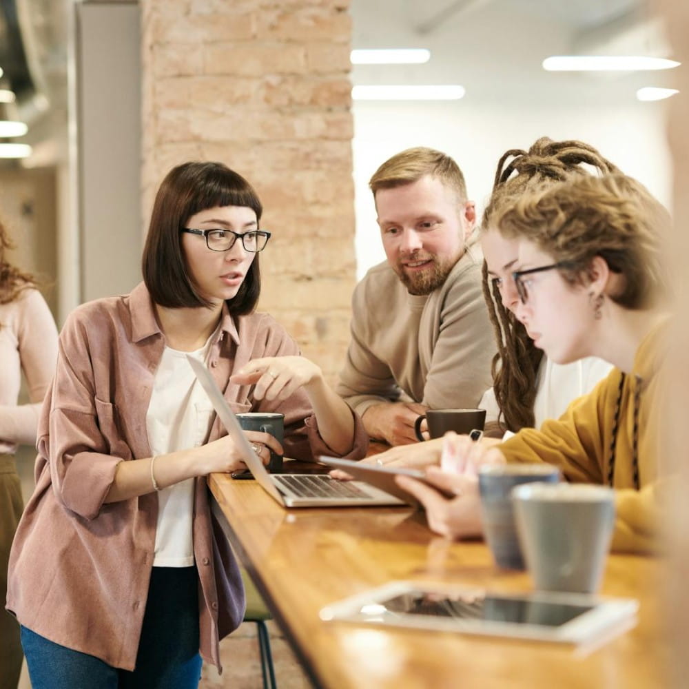 A group of people talking together while working on a laptop on a table, with coffee cups and an iPad in the foreground.
