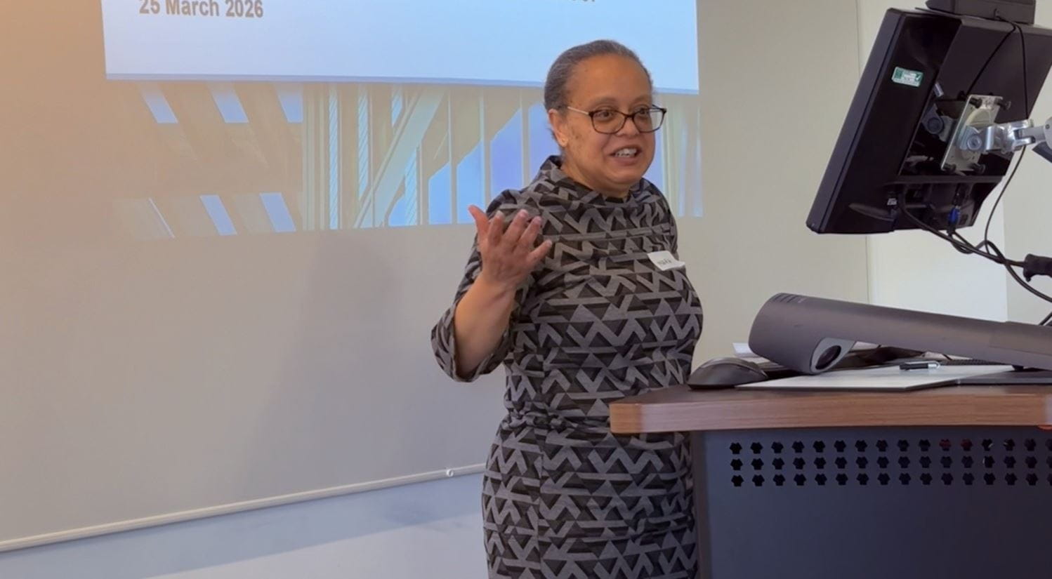 Dr Maria Hudson standing in front of a screen with talk slides projected on to it.