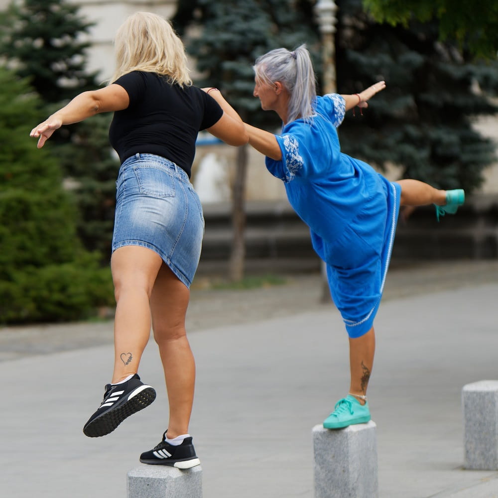 Two women standing on small pillars on one leg, their other leg out to the side.
