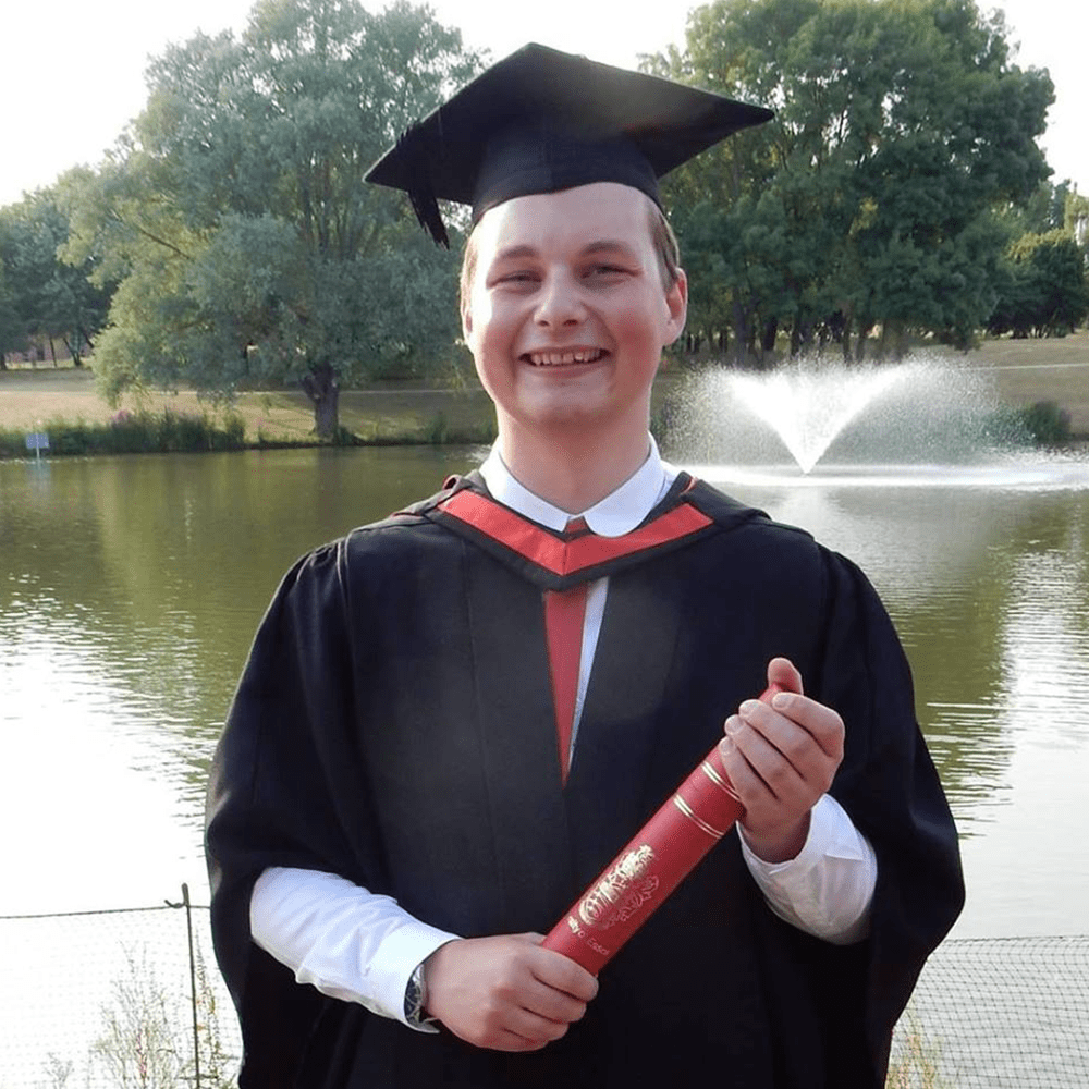 Perry at his graduation, wearing his robes and mortar board. He's smiling and holding his scroll in front of the famous Essex lakes.